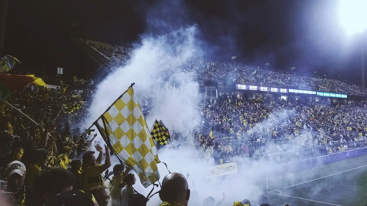 Fyldt fodboldtribune med fanflag og røg under nattebelysning